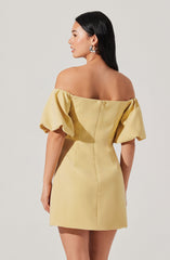 A woman is wearing a short, off-the-shoulder dress in a pale yellow color. The dress features voluminous puffed sleeves and a fitted silhouette with visible seam details. The back of the dress has a zipper closure, and she is accessorizing with large silver hoop earrings.
