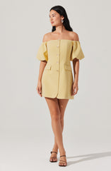 A woman is wearing a short, yellow, off-the-shoulder dress with puffy sleeves. The dress features large buttons down the front and two decorative flap pockets at the waist. She is also wearing strappy high-heeled sandals.