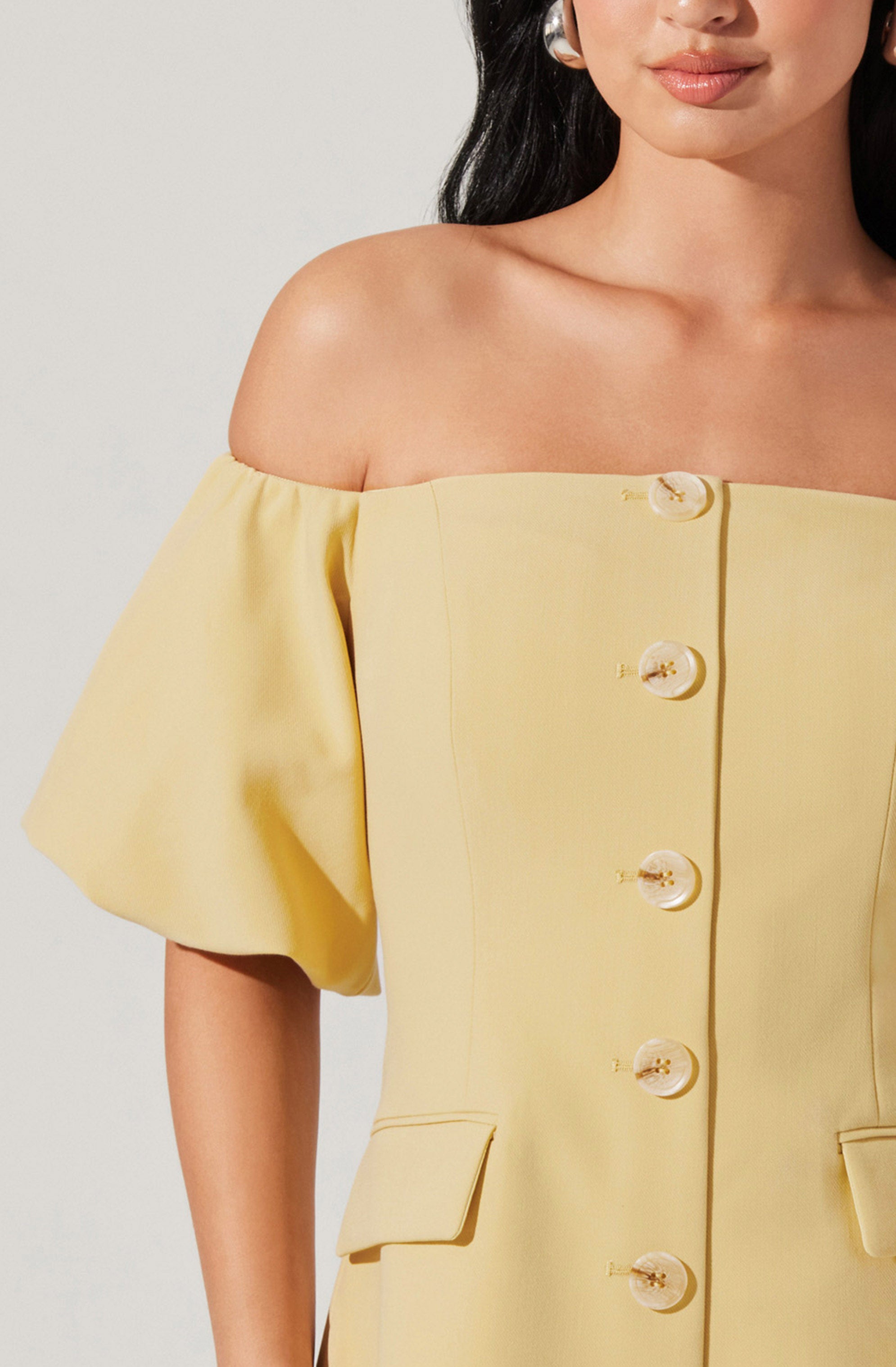 A woman is wearing a light yellow off-the-shoulder dress with large, puffy short sleeves and a row of large, round, pearl-like buttons down the front. The dress has a structured fit with small flap pockets near the waist.