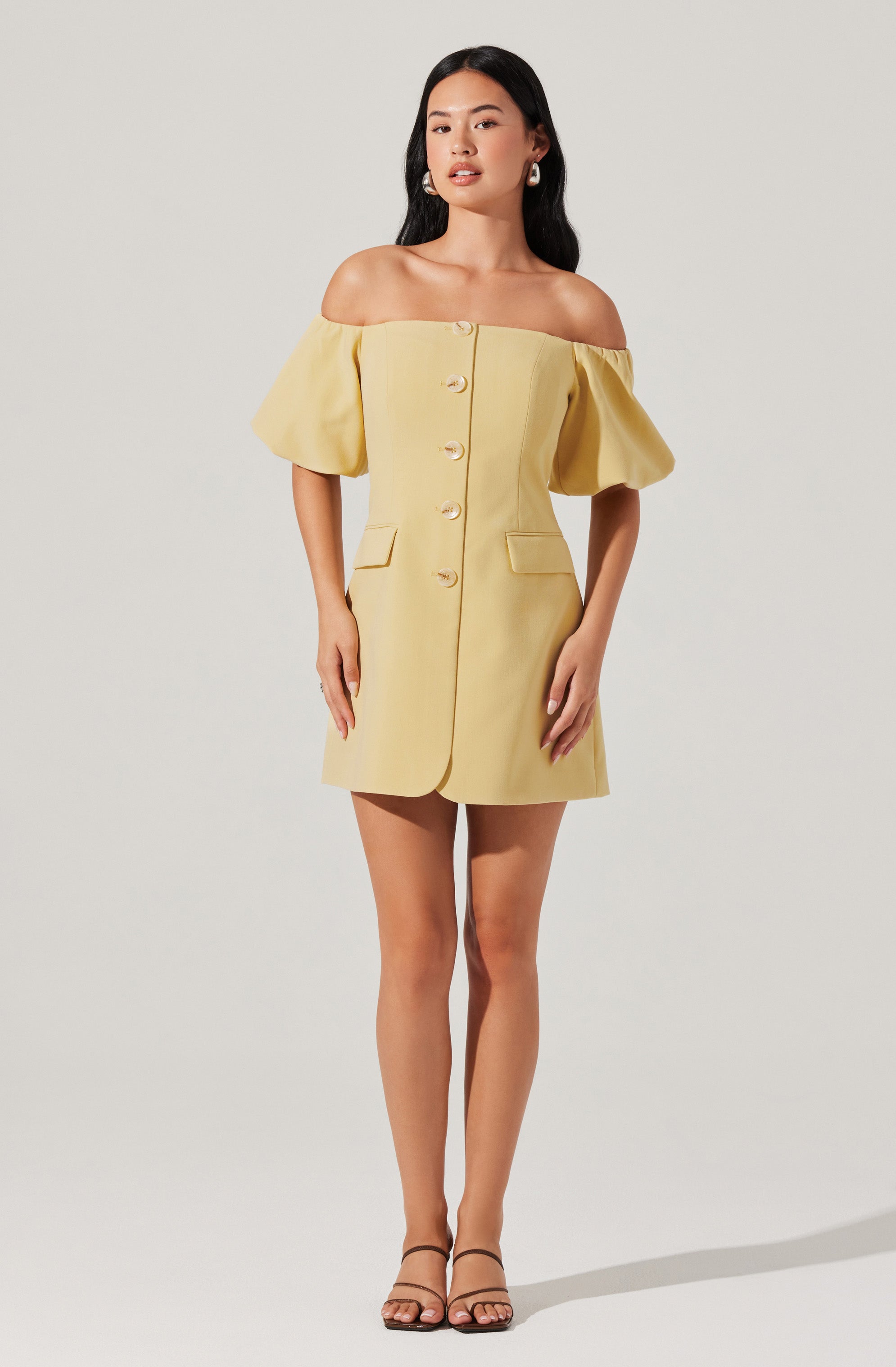 A woman is wearing a light yellow, off-the-shoulder dress with short, puffed sleeves. The dress features large buttons down the front and has two small pocket flaps at the hips. She is also wearing strappy, open-toe sandals and statement earrings.
