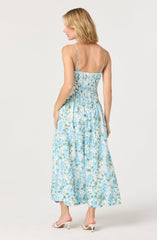 A woman is wearing a sleeveless, ankle-length dress with a smocked, fitted bodice and thin, adjustable spaghetti straps. The dress features a blue and white floral print, with the fabric gathered at the waist to create a full, flowing skirt. She is also wearing white heeled sandals. Her blonde hair is styled in a wavy, shoulder-length bob.