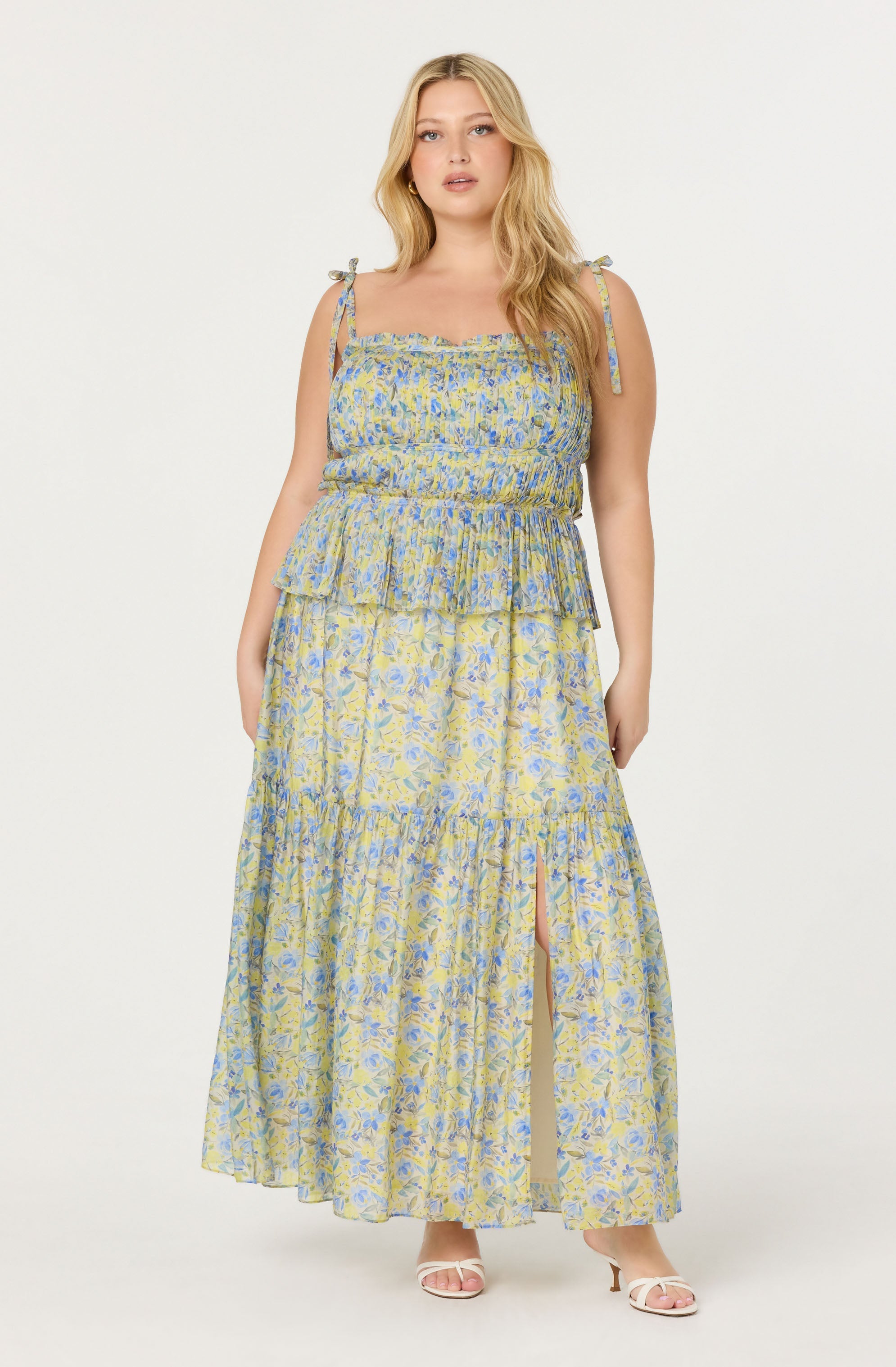 A woman is wearing a sleeveless, floor-length dress with a floral pattern in shades of yellow, blue, and green. The dress features adjustable tie straps on the shoulders, a ruffled neckline, and a tiered, flowing skirt with a front slit revealing her legs. She is also wearing open-toed high heel sandals.