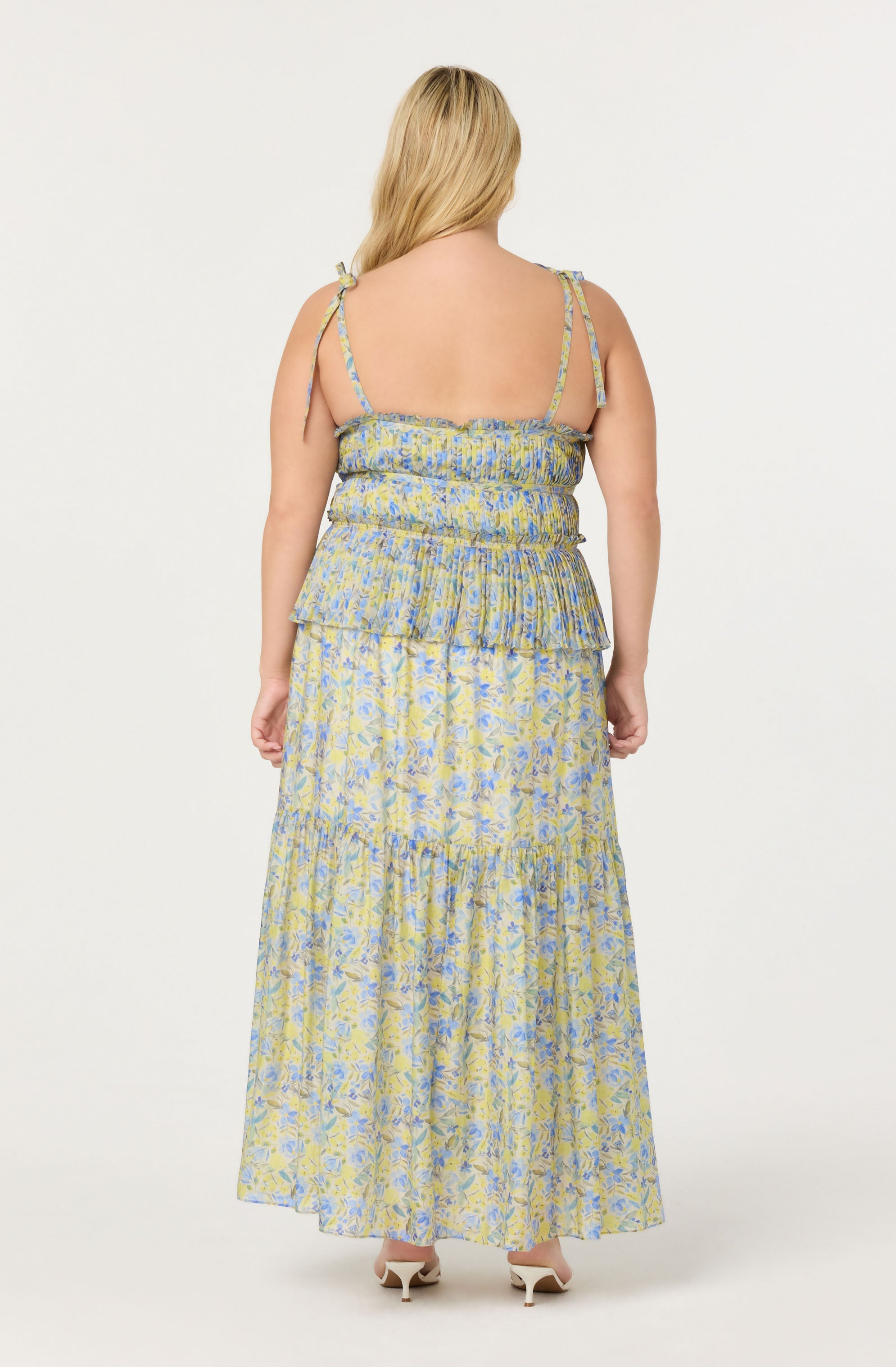 A woman is wearing a sleeveless, layered maxi dress with thin, adjustable spaghetti straps. The dress features a smocked bodice with a ruffled overlay and a floral pattern in shades of blue, yellow, and light green. She is also wearing open-toe, white high-heeled shoes.