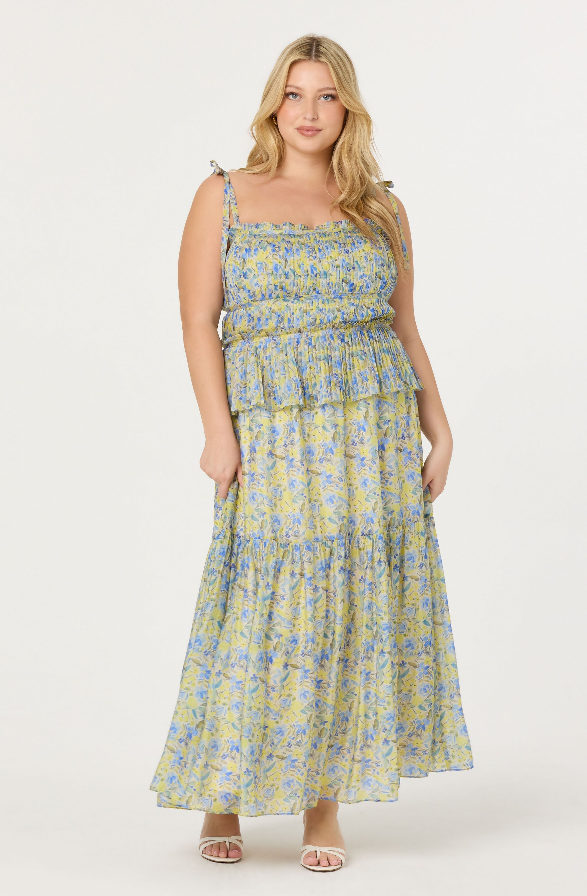 A woman is wearing a sleeveless, floral maxi dress with thin shoulder straps tied into bows. The dress features a smocked bodice and a tiered, flowing skirt, with a light-colored background and blue and yellow floral patterns. She is also wearing open-toe sandals.