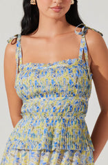 A woman is wearing a sleeveless dress with shoulder ties. The dress features a pastel-colored floral pattern in shades of blue, yellow, and white, with a smocked bodice and a tiered, ruffled skirt. The fabric appears lightweight and summery.