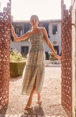 A woman is wearing a sleeveless, ankle-length dress with thin shoulder straps. The dress features a fitted bodice with a cinched waist and a flowing skirt with a slit up the front. The fabric has a floral pattern in shades of blue and yellow. She pairs the dress with open-toed high-heeled shoes.