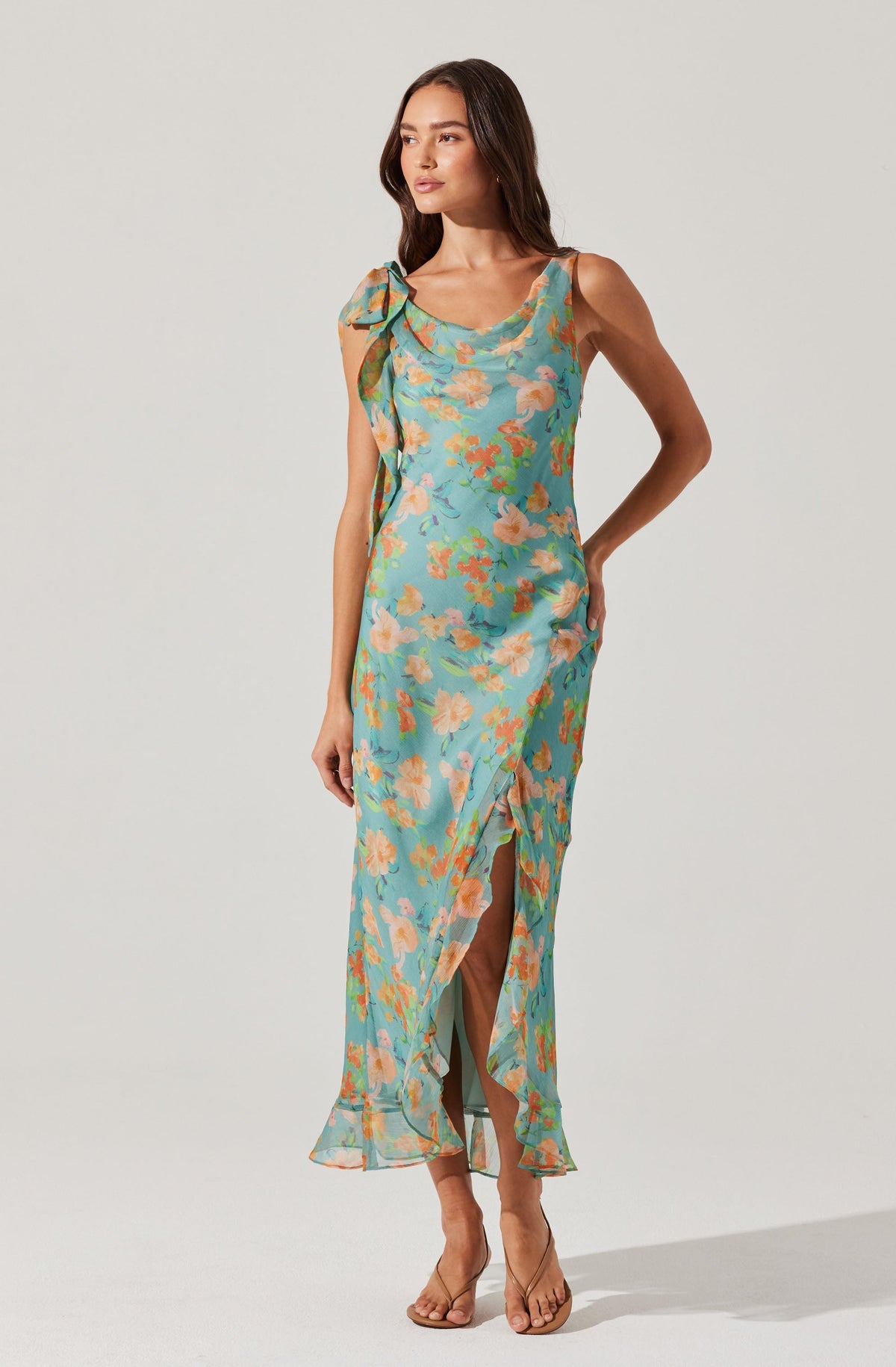 A woman is wearing a sleeveless, ankle-length dress with a floral pattern in shades of blue, peach, and green. The dress features a ruffled neckline, a slit at the front, and a large bow detail on one shoulder. She is also wearing nude-colored, open-toe high heels.