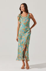 A woman is wearing a sleeveless, ankle-length dress with a floral pattern in shades of blue, peach, and green. The dress features a ruffled neckline, a slit at the front, and a large bow detail on one shoulder. She is also wearing nude-colored, open-toe high heels.