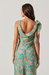 A woman is wearing a sleeveless dress with a floral pattern. The dress has a scoop neckline at the back and features a large, decorative bow or fabric detail on the left shoulder. The fabric is lightweight with a colorful print of orange and beige flowers with green leaves on a light blue background.