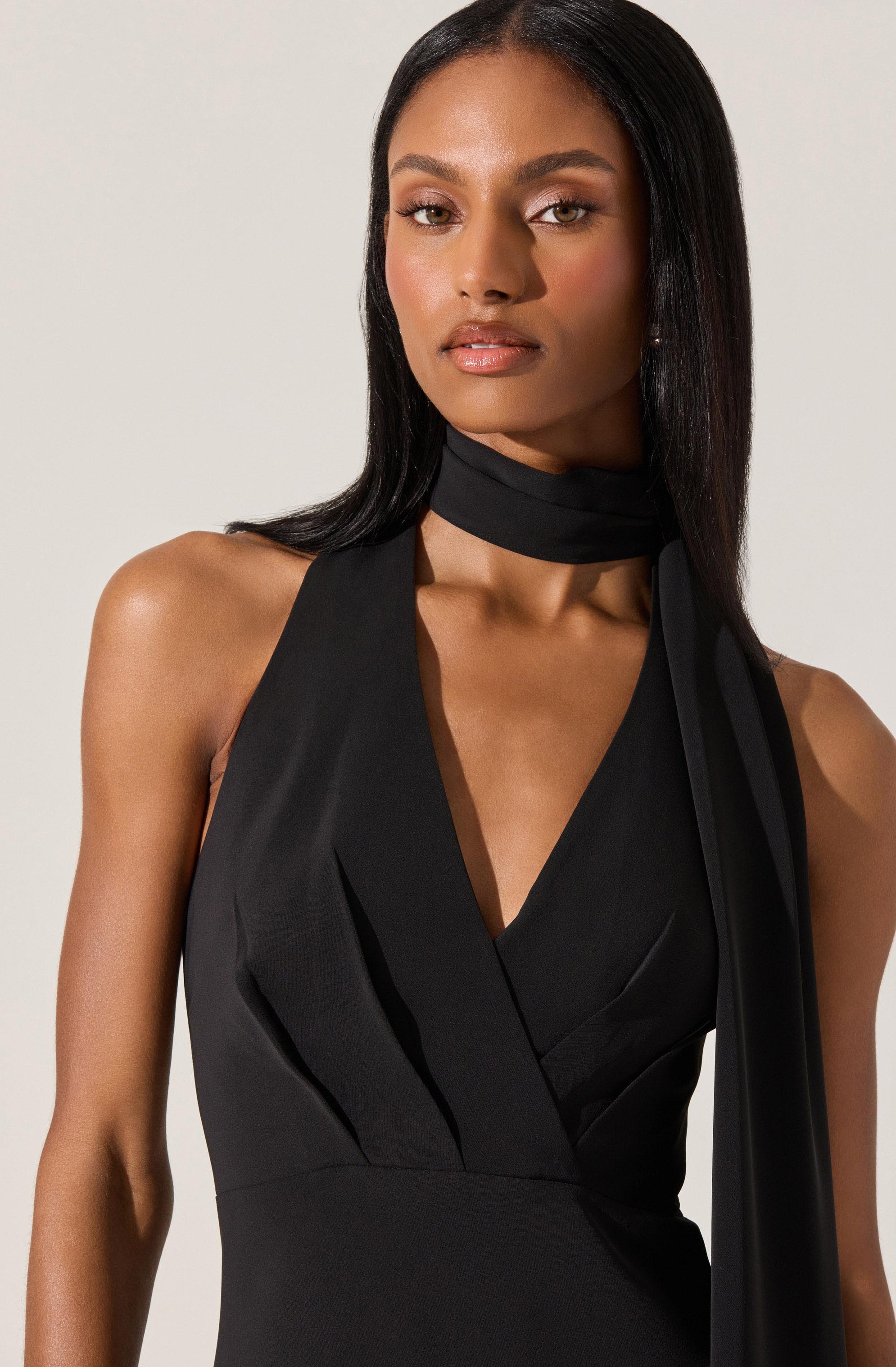 The image shows a woman wearing a sleeveless black dress with a deep V neckline and pleats on the bodice. She has a matching black scarf or necktie wrapped around her neck, hanging loosely over her shoulder. The overall style of the clothing is elegant and sophisticated.