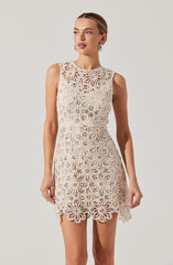 A woman is wearing a sleeveless, knee-length dress made of white lace with a floral, geometric pattern. The dress features a scalloped hemline and a fitted waist, with intricate openwork throughout the fabric.
