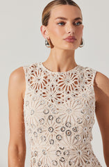 A woman is wearing a sleeveless, cream-colored crochet top featuring a floral pattern with circular and radiating petal designs. The top has a high neckline and intricate openwork details. She accessorizes with large, shiny silver hoop earrings.