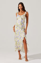 A woman is wearing a sleeveless, ankle-length dress with a floral pattern in shades of purple, green, and white. The dress features ruffled shoulder straps, a ruffled neckline, and a layered, asymmetrical hem with a high slit on one side. She is also wearing open-toed, strappy high-heeled sandals.