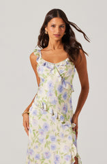 A woman is wearing a sleeveless, ankle-length dress with a floral print in shades of light purple, green, and cream. The dress features ruffles along the neckline, shoulder straps, and down the side, creating a layered, textured look.