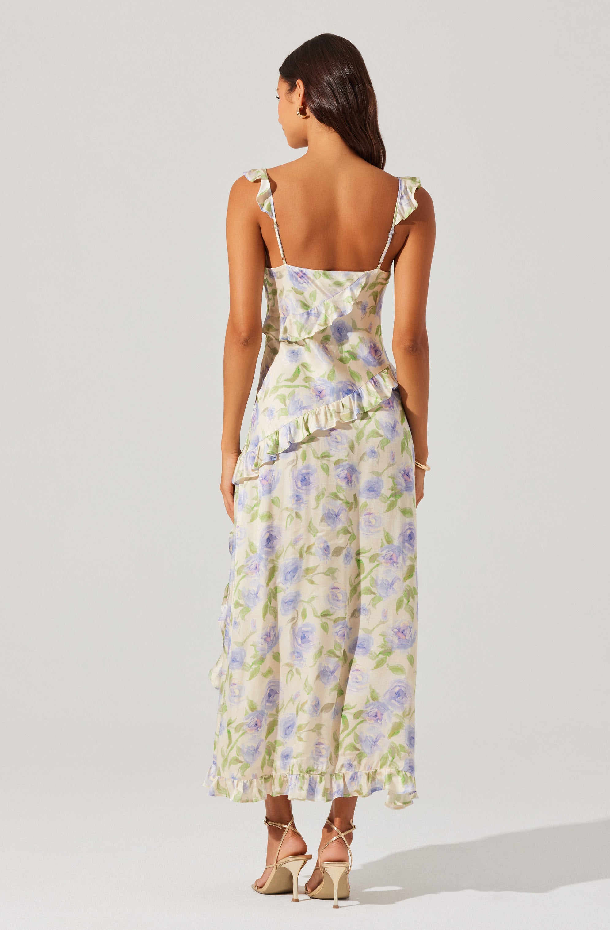 A woman is wearing a sleeveless, ankle-length dress with ruffled shoulder straps and a ruffled overlay at the waist. The dress features a light pastel floral print with shades of purple, green, and cream. She is also wearing beige high-heeled sandals.