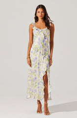 The woman is wearing a sleeveless, ankle-length dress with a light-colored background and a pastel floral print in shades of purple, green, and blue. The dress features ruffle details along the neckline, armholes, and hem, as well as a high slit on one side. She is also wearing nude strappy high-heeled sandals.