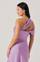 A woman wearing a sleeveless, light purple satin dress with a one-shoulder neckline. The dress features a draped fabric detail on the shoulder, which extends across the top of the dress. The fabric has a smooth, shiny texture, and the dress fits snugly around her waist and hips. She is accessorized with large, silver hoop earrings and has long, straight black hair.