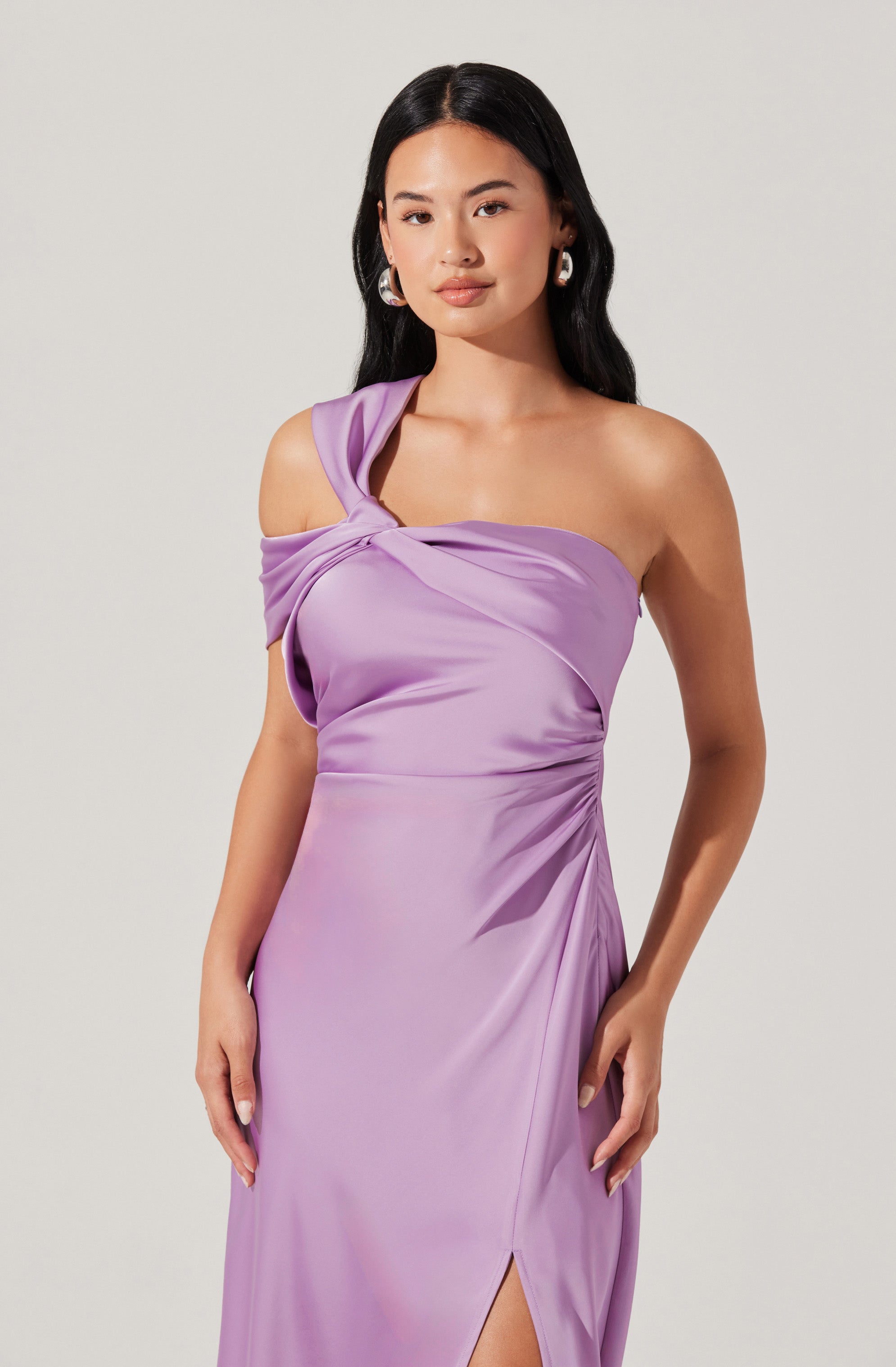A woman is wearing a sleeveless, lavender satin dress with an off-the-shoulder design on one side. The dress features a twisted knot detail at the shoulder and a thigh-high slit on the side.
