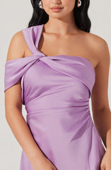 A woman is wearing a lavender satin dress featuring a one-shoulder design with a large, draped bow on the shoulder. The fabric is smooth and shiny, with gentle ruching at the waist.