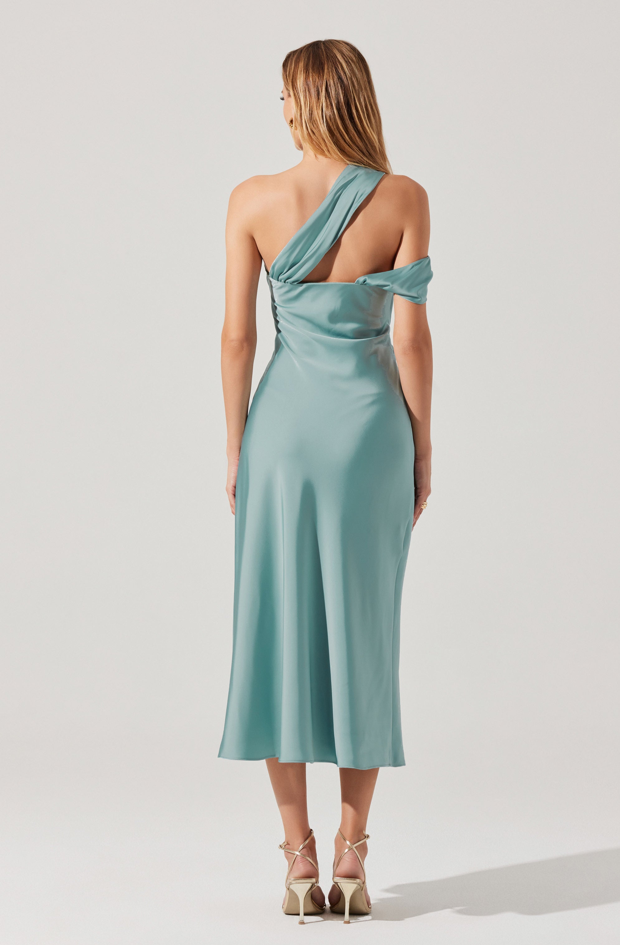 A woman is wearing a satin, light teal, one-shoulder dress with a draped design. The dress features an asymmetrical neckline with a wide strap on one shoulder and a loose fabric detail on the opposite side. The length of the dress reaches mid-calf. She is also wearing beige high-heeled sandals with thin straps.