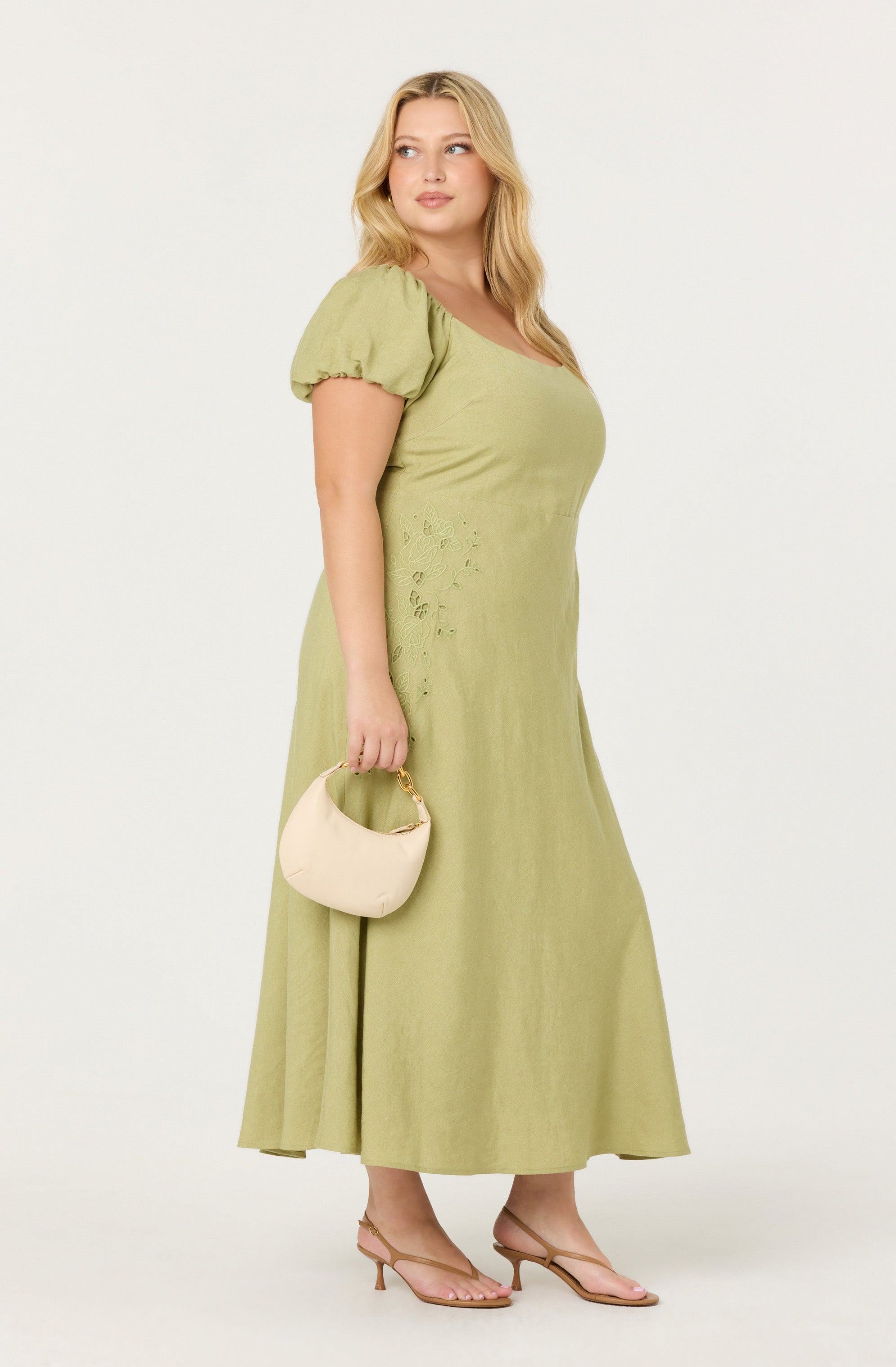 A woman is wearing a light green, ankle-length dress with puffed short sleeves and subtle embroidery or eyelet detailing on the side. She is holding a small, light beige handbag with a gold handle and is wearing beige open-toe high-heeled sandals.