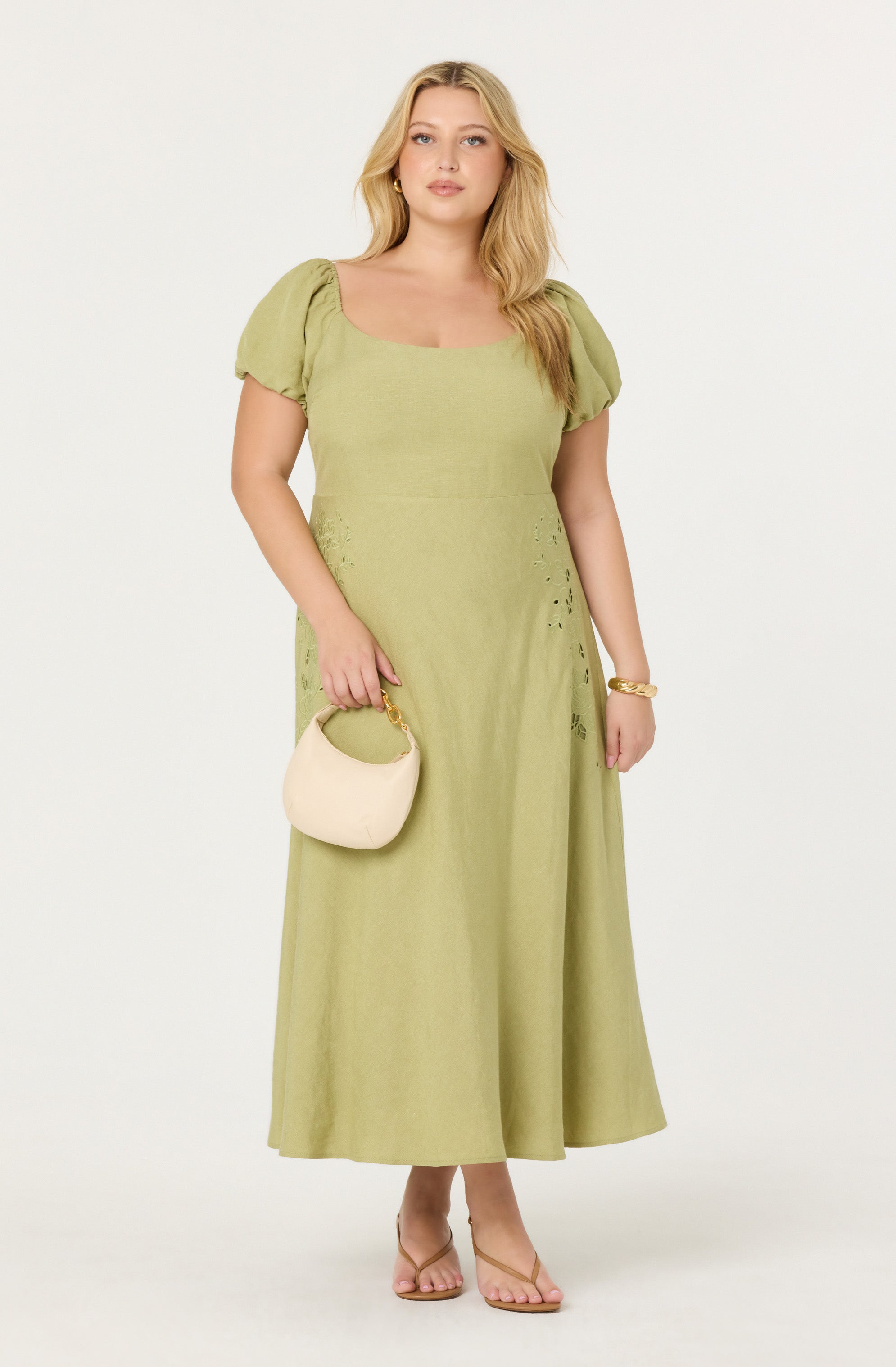 A woman is wearing a light green, ankle-length dress with puffed short sleeves and a square neckline. The dress features small cut-out details on the sides near the waist. She is holding a small, white handbag with a gold handle. She is also wearing a gold bracelet on her right wrist and beige open-toe high-heeled sandals.