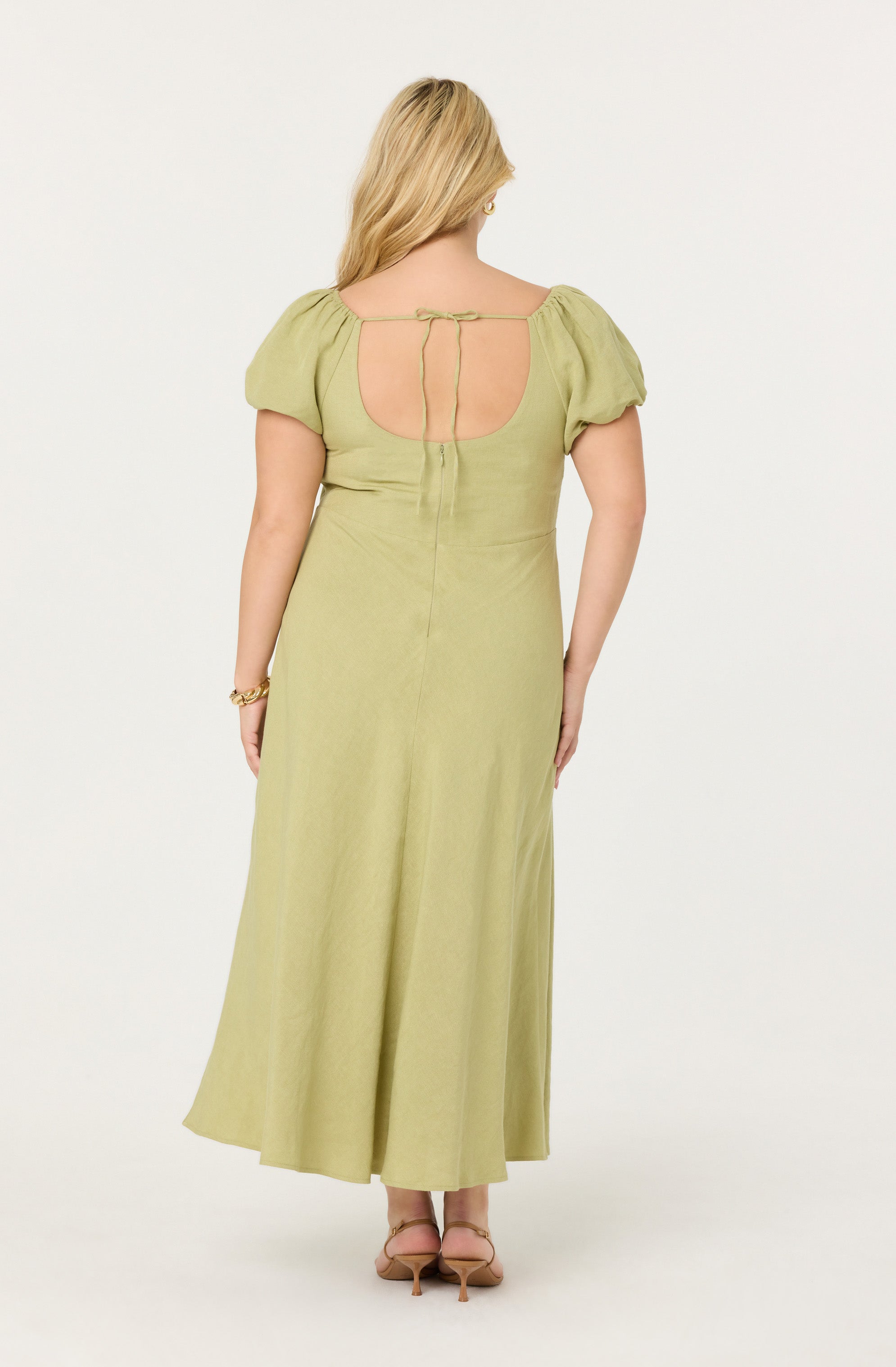 A woman is wearing a light green, floor-length dress with short puffed sleeves. The back of the dress features a large open oval cutout and a thin tie closure at the top. She is also wearing strappy high-heeled sandals and a bracelet on her right wrist.