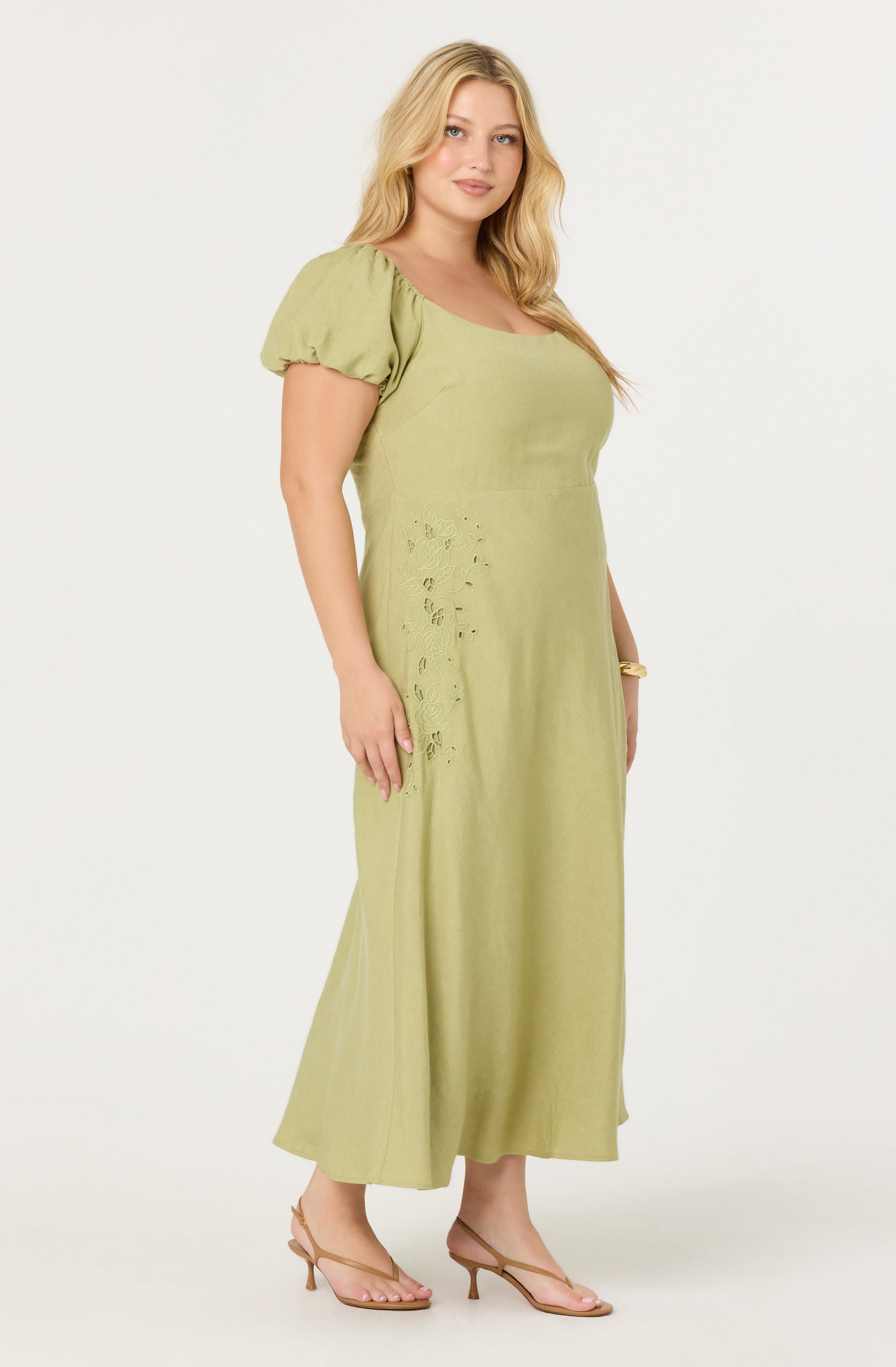 A woman is wearing a light green maxi dress with short, puffed sleeves. The dress features subtle eyelet embroidery details on the side near the waist. She is accessorizing with a minimalist bracelet and wearing nude-colored heeled sandals.