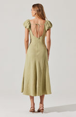 A woman is wearing a light green, ankle-length dress with puffed short sleeves. The dress features a deep, open back with a tie detail at the top and a visible zipper running down the center back. She is also wearing nude-colored high-heeled sandals.