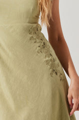 A close-up of a light green dress made of smooth fabric featuring intricate floral embroidery with roses and leaves along the side. The embroidery is detailed with cut-out sections, adding texture to the design. The dress is fitted at the waist, and a portion of the person's arm and hand are visible, with long, wavy hair seen in the background.