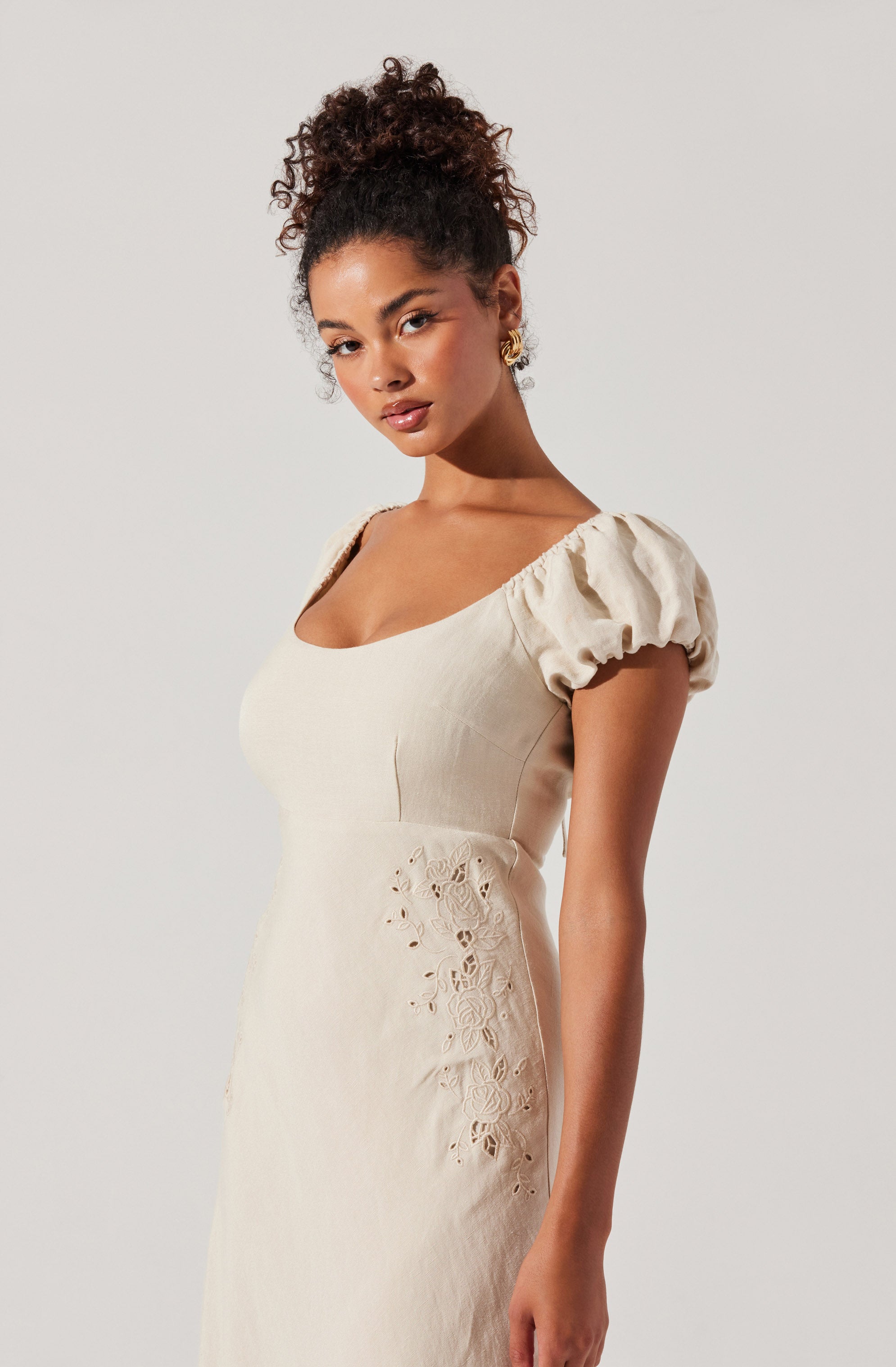 A woman is wearing a light beige dress with puffed short sleeves and a fitted bodice. The dress features embroidered cutout details along the side.