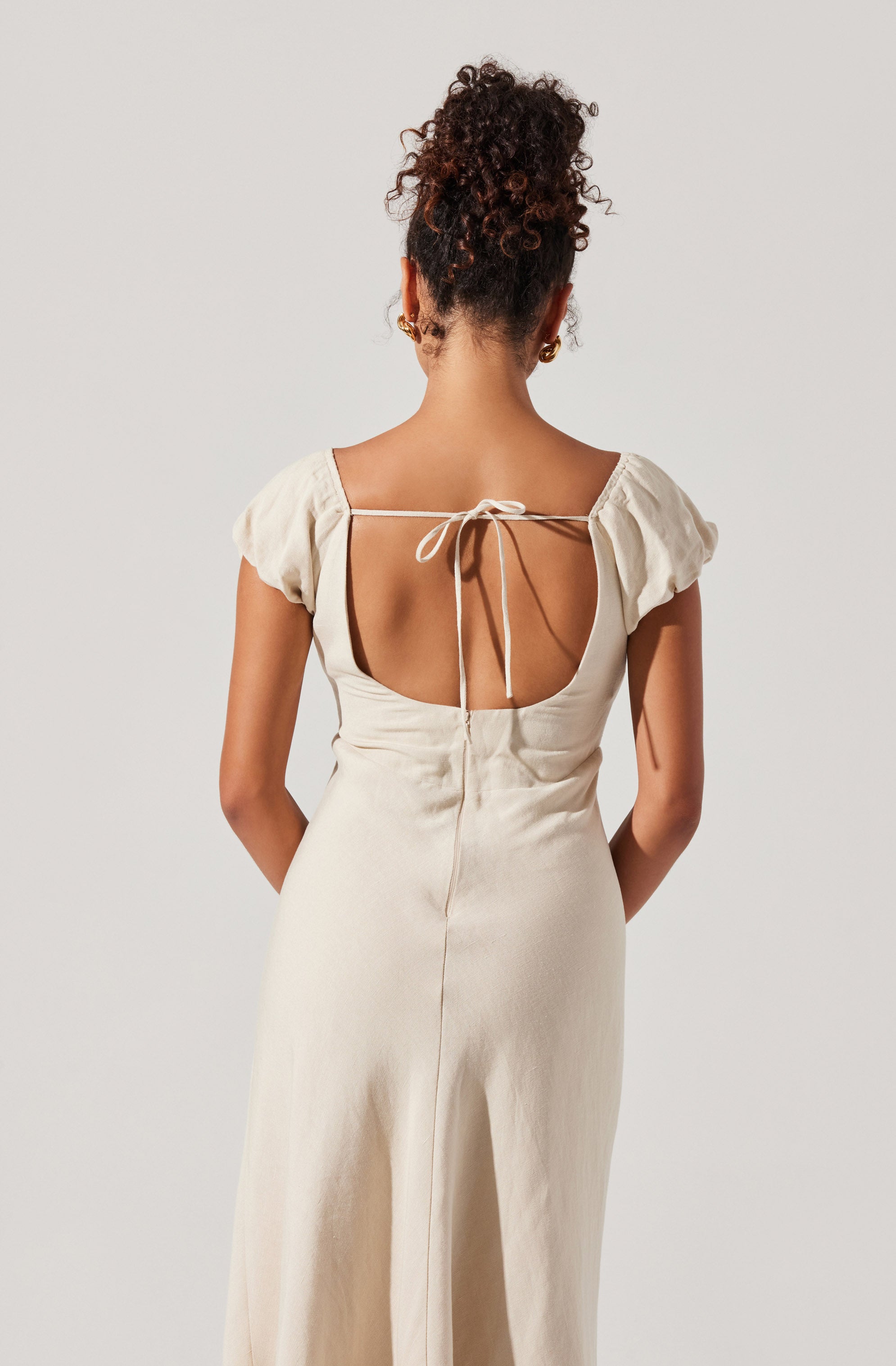 A woman is wearing a cream-colored, satin-like dress with short, puffed sleeves. The dress has a low open back with a thin strap tied in a bow at the top. The material appears smooth and shiny, and the dress extends down past her hips.