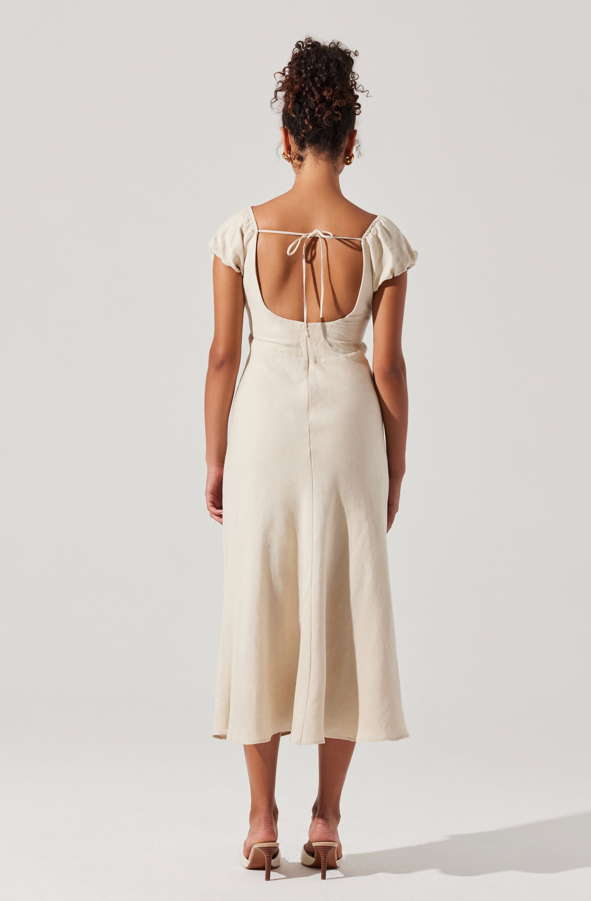 A woman is wearing a cream-colored, sleeveless, ankle-length dress with a fitted waist and a front slit. The dress features a low back with a tie-up detail at the shoulders. She is also wearing a pair of beige high-heeled sandals.