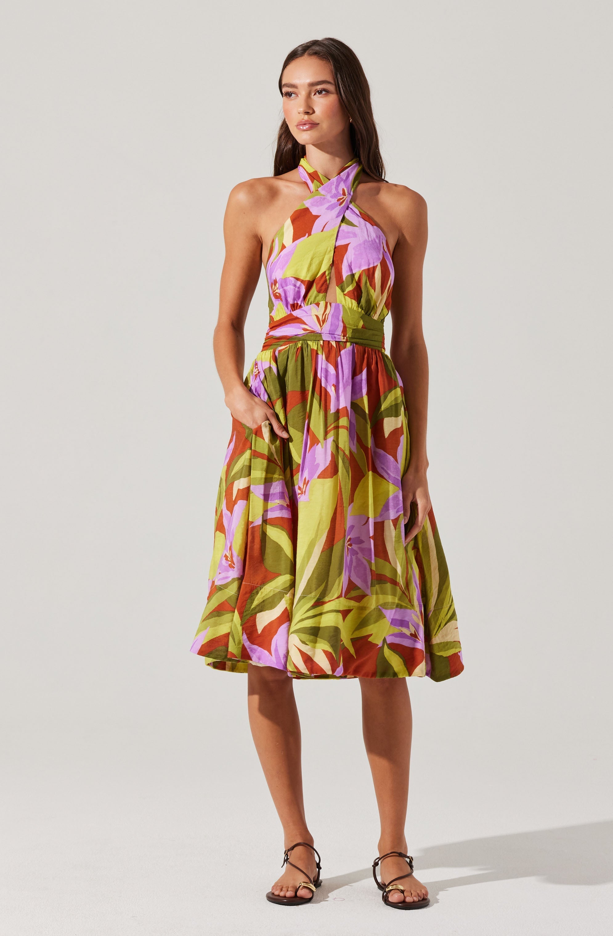 A woman is wearing a sleeveless, halter-neck dress with a colorful, large floral pattern in shades of purple, pink, green, and brown. The dress has a fitted bodice and a full, knee-length skirt.