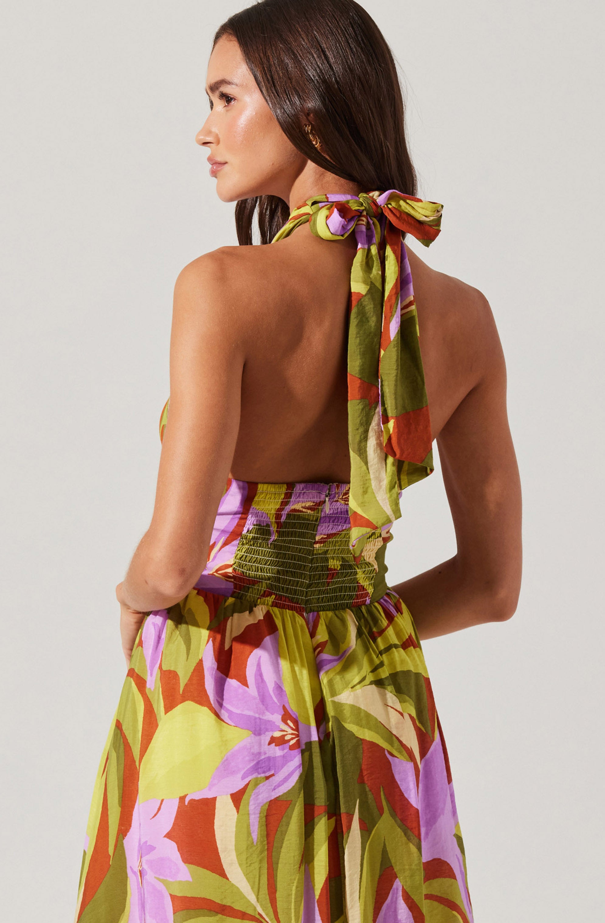 A woman is wearing a colorful, sleeveless, backless dress with a halter tie strap around her neck. The dress features a vibrant floral pattern in shades of purple, pink, orange, and green. The fabric appears lightweight and flowy.