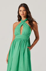 A woman wearing a sleeveless, bright green halter dress with a crisscross neckline that features a keyhole opening at the chest. The dress has a gathered, elastic waistband and a full, flowing skirt. She has her right hand in her pocket and her left arm resting at her side.