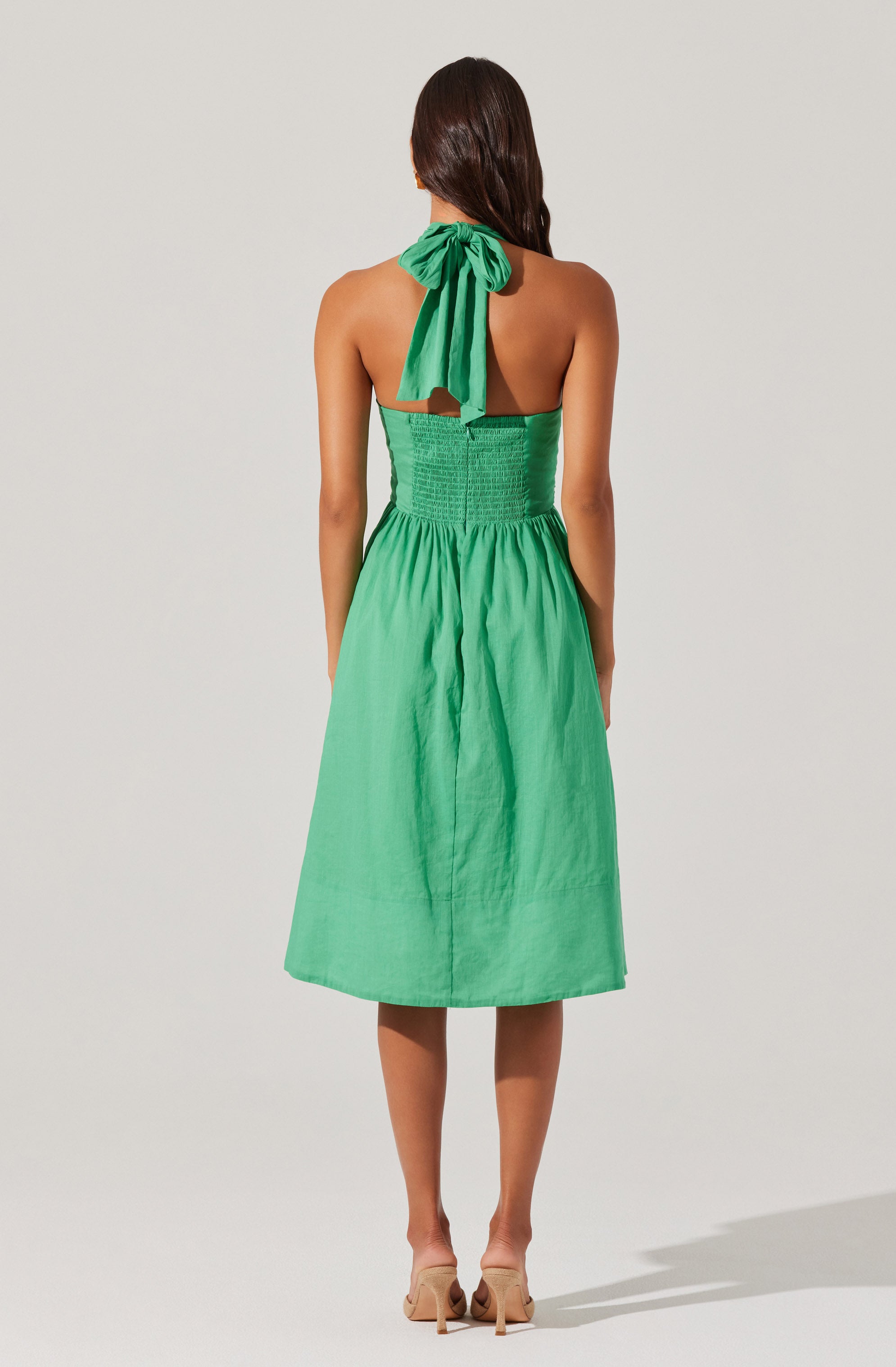 The dress is sleeveless with a halter neckline tied at the neck with a large bow. The bodice is fitted with shirred, textured fabric, and the skirt is flowy and knee-length, made of a lightweight, solid green fabric. The model is wearing nude high-heeled shoes.