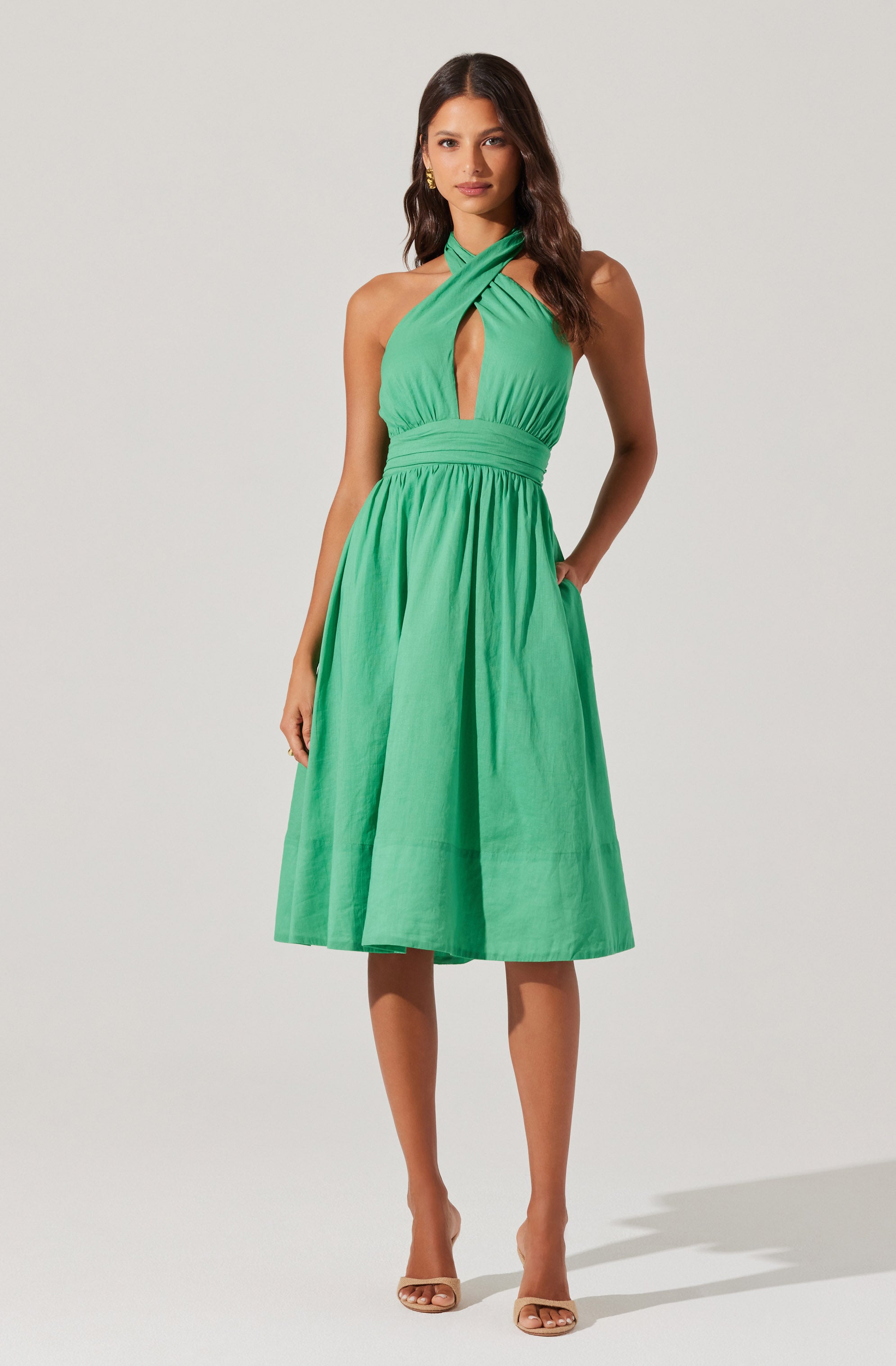 A woman is wearing a sleeveless, halter dress in a bright green color. The dress features a criss-cross halter neckline with a keyhole cutout in the front. It has a fitted waistband and a full, knee-length A-line skirt. The fabric appears lightweight and slightly textured. She is also wearing nude open-toe high heels.