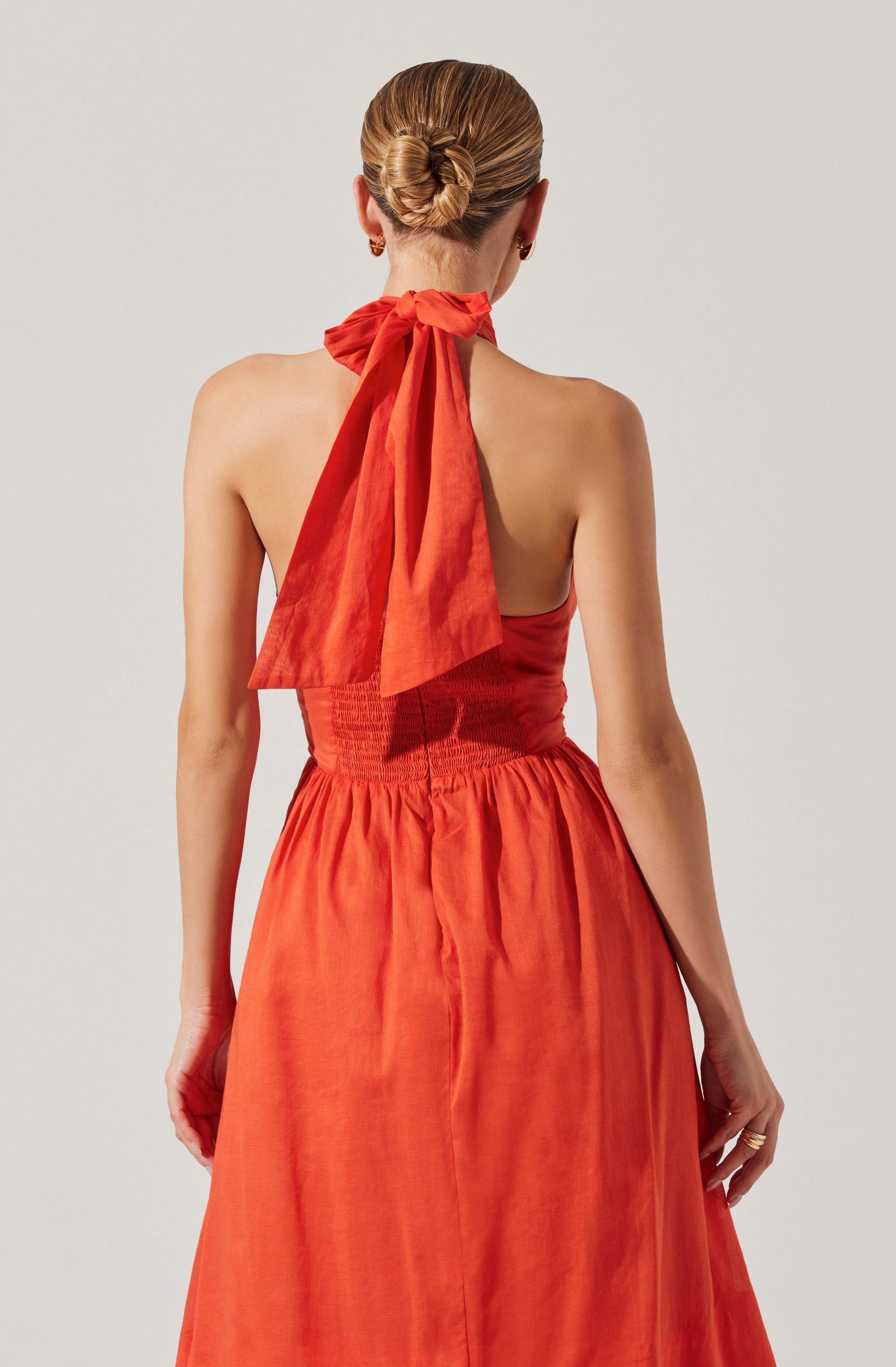 The image shows a woman from behind wearing a sleeveless, strapless dress in a coral or burnt orange color. The dress has a fitted, ruched bodice and a gathered, flowing skirt. She has her hair styled in a neat bun and is accessorized with small earrings and a ring. A notable feature is the large, matching orange bow tied around her neck, which drapes down her back.