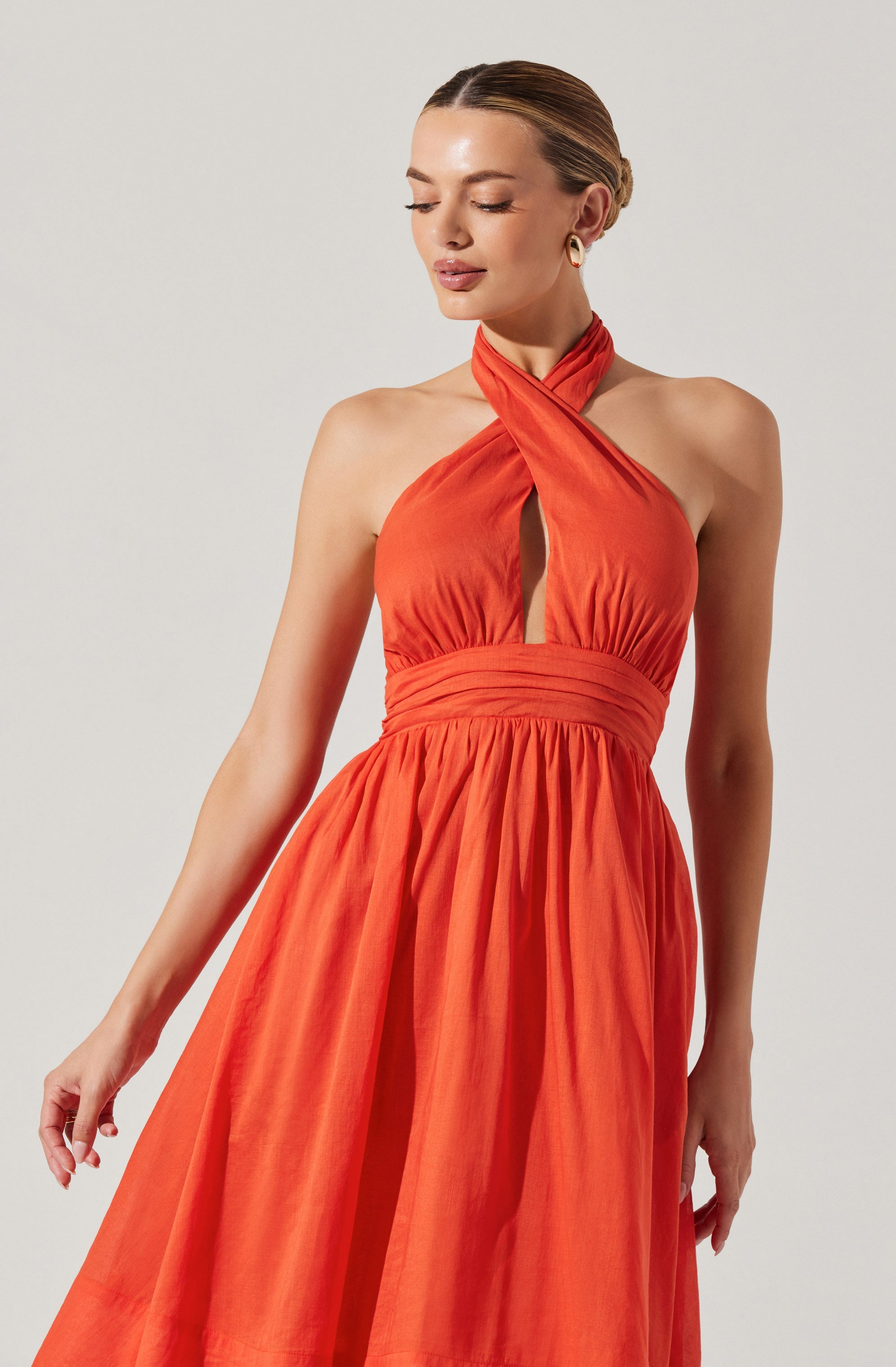 A woman is wearing a sleeveless, halter-style orange dress with a gathered waistband. The dress features a high neckline that twists around the neck, with a keyhole cutout in the front. The skirt is full and gathered, creating a flowing silhouette. The fabric appears to be lightweight and slightly textured.