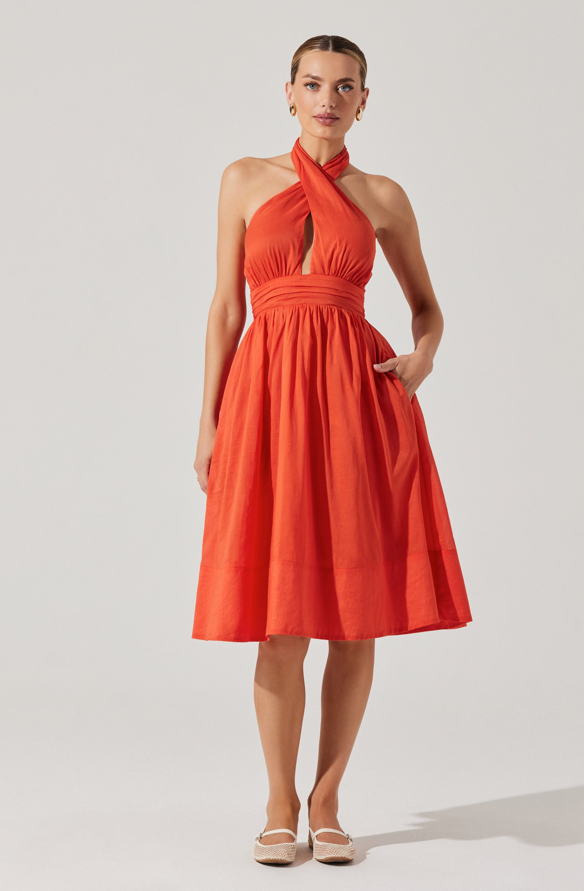 A woman is wearing a sleeveless, halter-neck dress in a bright orange color. The dress features a fitted bodice with a keyhole cutout at the front and an elastic waistband. The skirt is knee-length, full, and gathered, creating a voluminous silhouette. She has her right hand in a pocket on the side of the skirt and is wearing light-colored, closed-toe shoes with a strap.