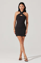 A woman is wearing a fitted black halter dress with horizontal ruched detailing throughout. The dress features a crossed strap design around the neck and a small cutout detail just below the neckline. She is accessorized with gold hoop earrings, gold bracelets, and rings, and is wearing black high-heeled sandals.