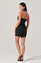 A woman is wearing a strapless black mini dress with ruching details throughout. She accessorizes with gold hoop earrings and a gold bracelet. She is also wearing black high-heeled sandals.
