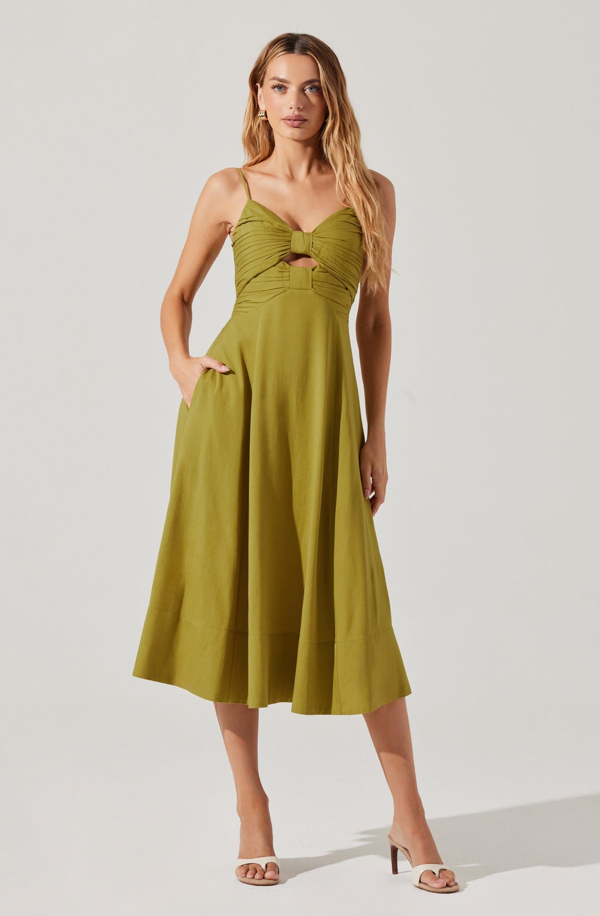 A woman is wearing a sleeveless, olive-green dress with spaghetti straps. The dress has a fitted bodice with a large, knotted detail at the center of the bust, creating a keyhole cutout below the knot. The skirt is full and flares out to mid-calf length, with a wide hem at the bottom. She completes the look with beige, open-toe high-heeled sandals.