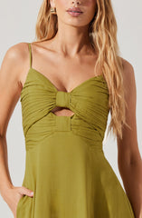 A woman is wearing a sleeveless, olive green dress with thin spaghetti straps. The dress features gathered fabric around the bust area with a large bow detail and a small cutout just below the bow. The skirt of the dress flows loosely, and she has one hand in her pocket. She is accessorized with gold hoop earrings.