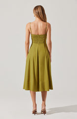 A woman is wearing a sleeveless, olive-green dress with thin spaghetti straps. The dress features a fitted, smocked bodice and a full, pleated skirt that falls to mid-calf length. She is also wearing nude-colored high-heeled sandals.