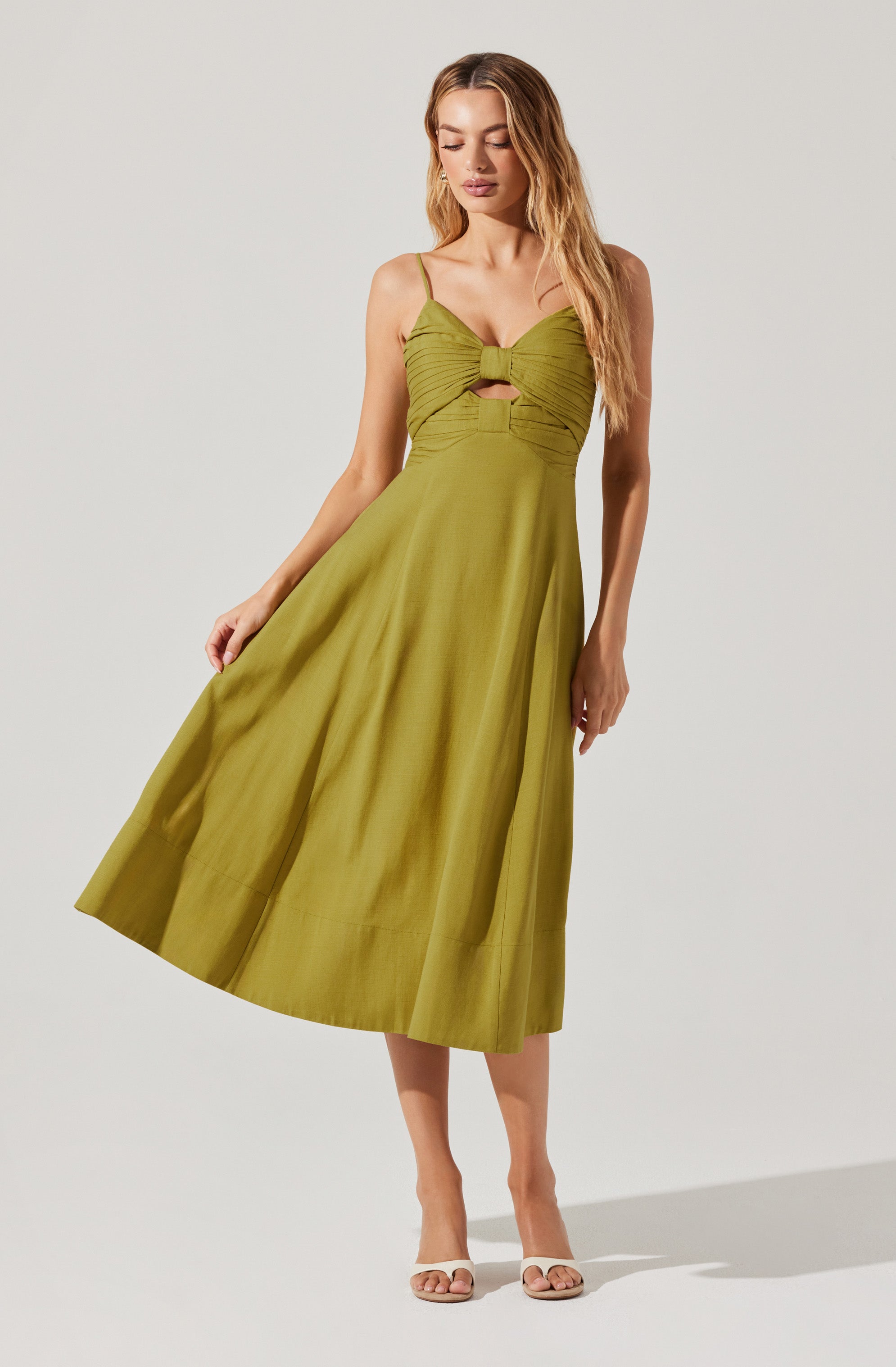 A woman is wearing a sleeveless, olive green dress with thin spaghetti straps. The bodice features a gathered, ruched design with a knotted detail at the center and a small cutout just below the knot. The skirt is full and midi-length, flowing gracefully to the mid-calf. She is also wearing light-colored open-toe high heels.