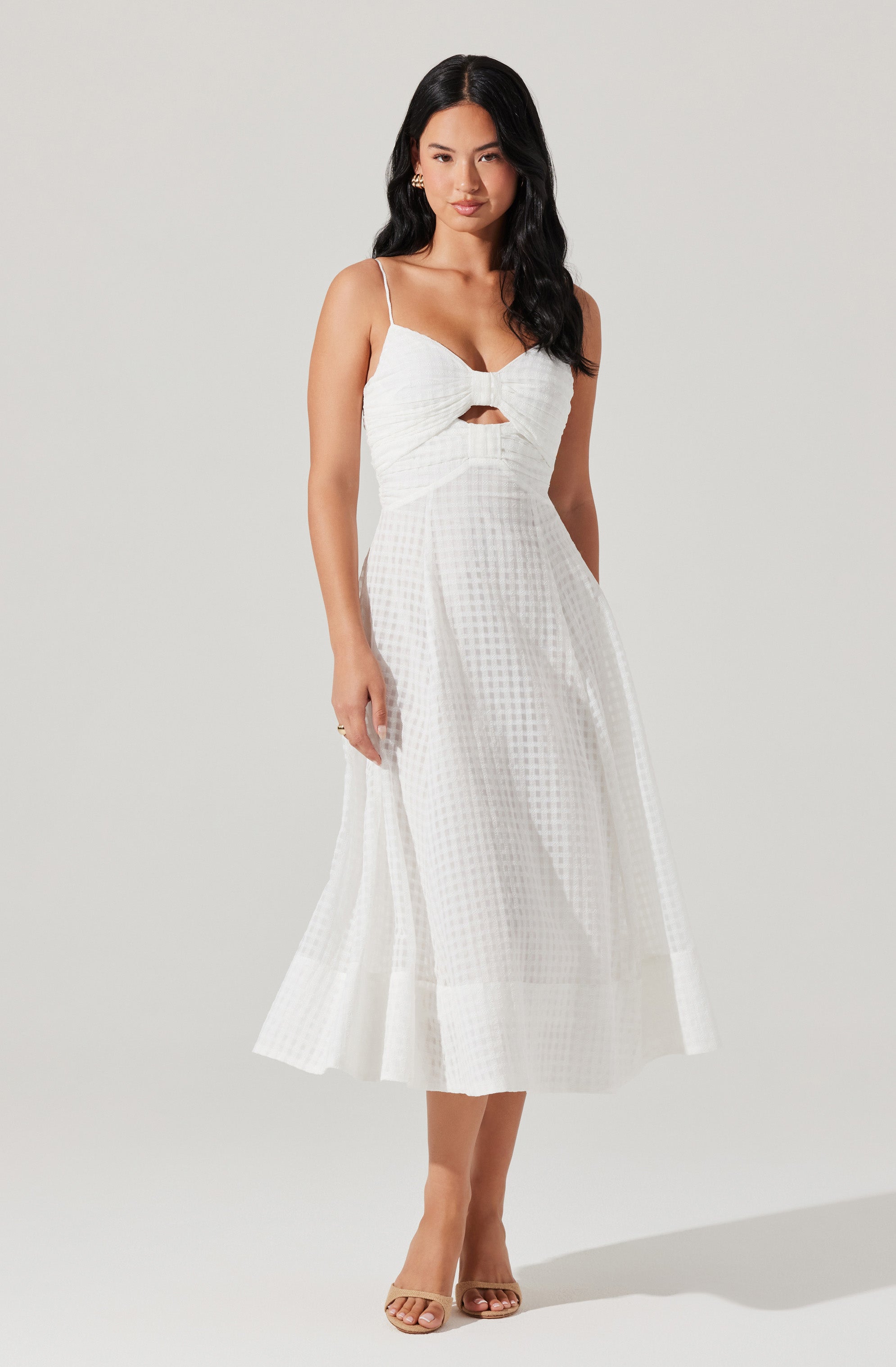 A woman is wearing a white, sleeveless dress with spaghetti straps. The dress features a fitted bodice with a ruched, bow-like detail at the chest and a small cutout below the bow. The skirt is high-waisted, flowing, and reaches mid-calf, with a subtle checkered pattern and a wide, pleated hemline.