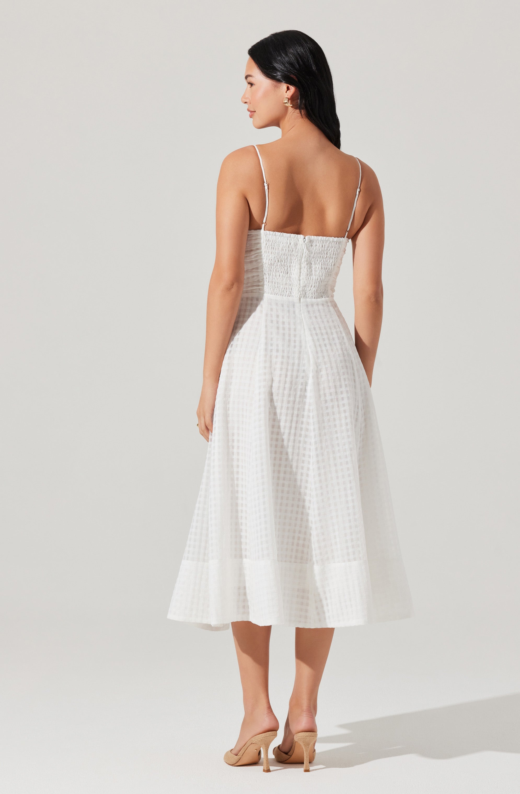 A woman is wearing a white dress with thin spaghetti straps, a fitted bodice with a smocked design, and a full, mid-length skirt made of a textured fabric with a subtle checkered pattern. She pairs the dress with nude high-heeled shoes.
