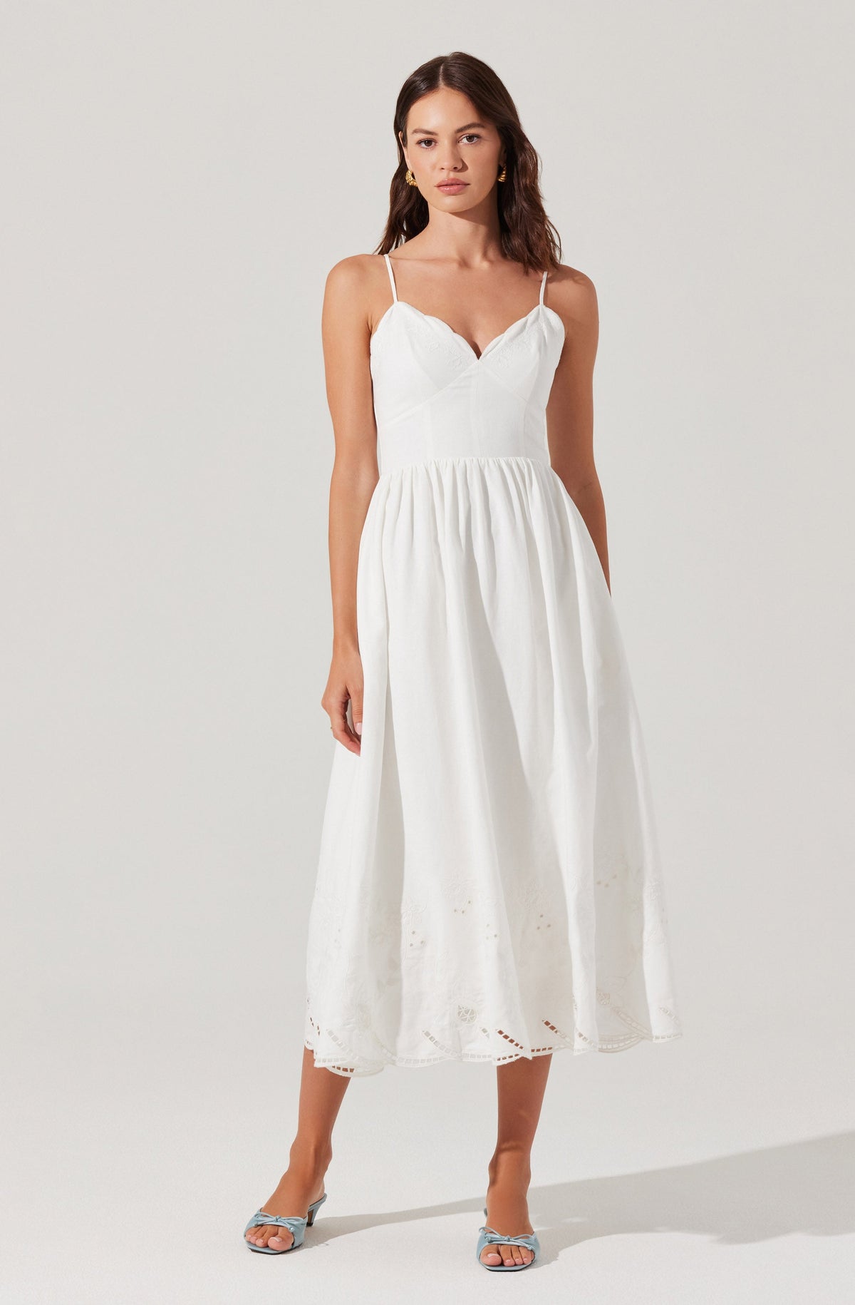 A woman is wearing a white, sleeveless, ankle-length dress with thin spaghetti straps. The dress has a fitted bodice with a V-shaped neckline and a gathered waist that flows into a full skirt. The skirt features eyelet embroidery near the hem, adding a delicate decorative detail. She is also wearing open-toed, strappy high-heeled sandals in a light color.